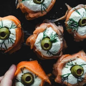 A hand holding a plate of smoked salmon rolls filled with cream cheese, topped with an olive slice and garnished with dill—perfect for those seeking one-click favorite recipes. The rolls are elegantly arranged in a circular pattern on a sleek black plate.