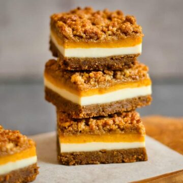 Three stacked dessert bars with layers of crumbly brown topping, orange filling, creamy white middle, and a brown base. They are placed on a wooden board with a grey background. Another bar is partially visible on the left.