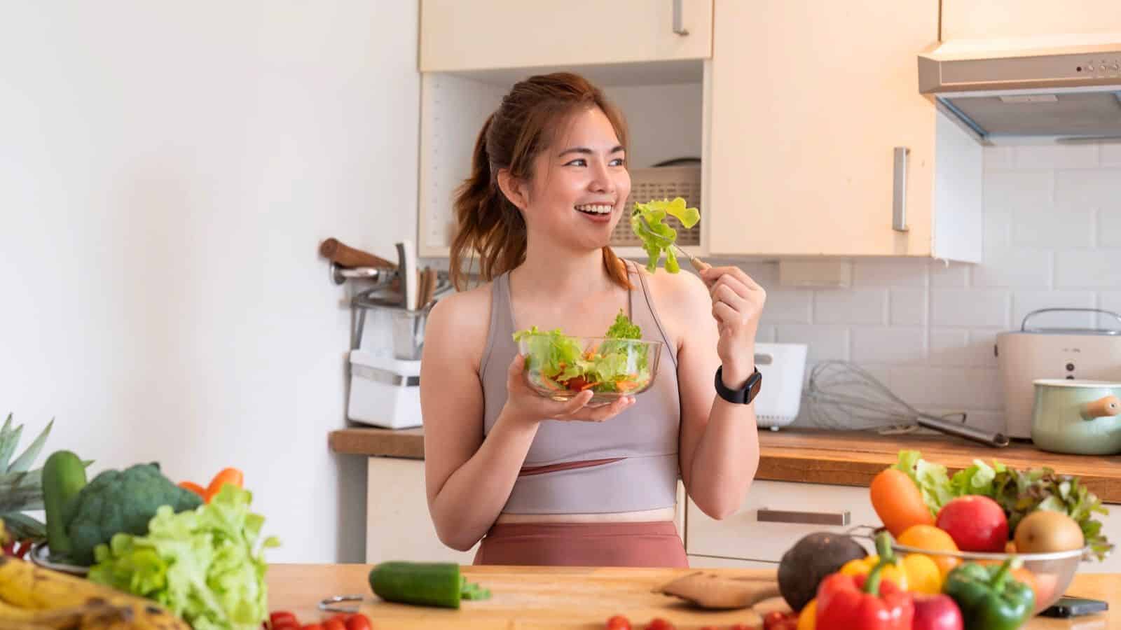 A woman in workout attire is standing in a kitchen, holding a bowl of salad with leafy greens. She is smiling and holding a piece of lettuce. The counter is filled with various fruits and vegetables.