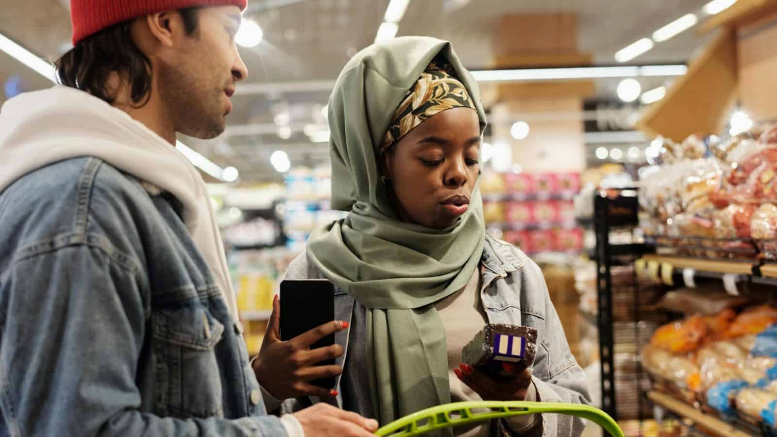 A man and a woman are shopping in a grocery store. The woman is wearing a headscarf and holding an avocado and a phone, while the man is wearing a red beanie. They are standing in front of shelves with bread and other products.