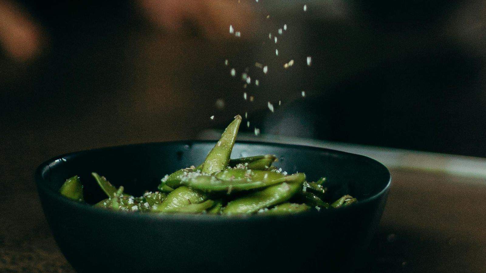 A black bowl filled with edamame pods is placed on a dark surface. Salt is being sprinkled over the edamame from above, with some grains visible in mid-air. The background is blurred and out of focus.