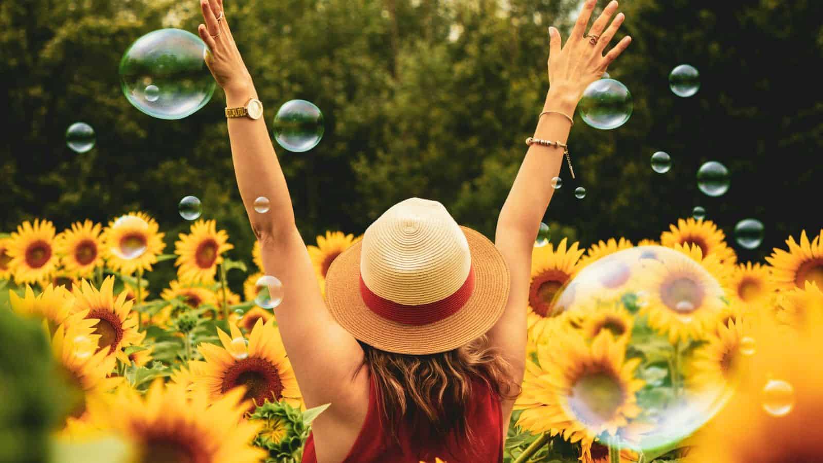 A person stands in a sunflower field with arms raised, wearing a straw hat and a sleeveless top. Bubbles float around them. The background consists of lush green trees under a lightly cloudy sky.