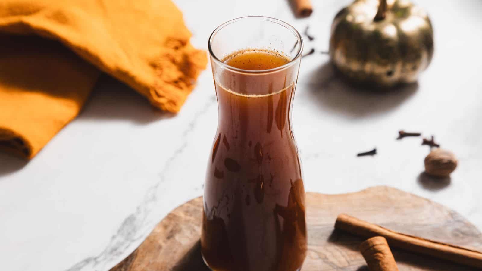 A glass carafe filled with dark brown apple cider sits on a wooden surface, offering refreshing sips enhanced by cinnamon sticks, cloves, and whole nutmeg. In the background, an orange cloth and a small decorative pumpkin create an inviting atmosphere for any day.