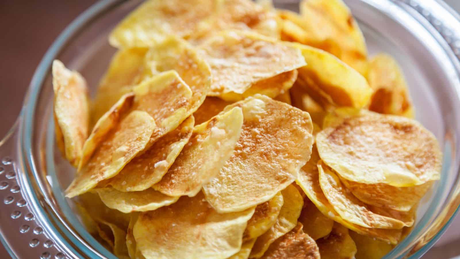 A clear glass bowl filled with golden brown potato chips sits on a surface. The chips are thinly sliced and appear crispy, with a light sheen on the surface, indicating they are freshly prepared or fried.