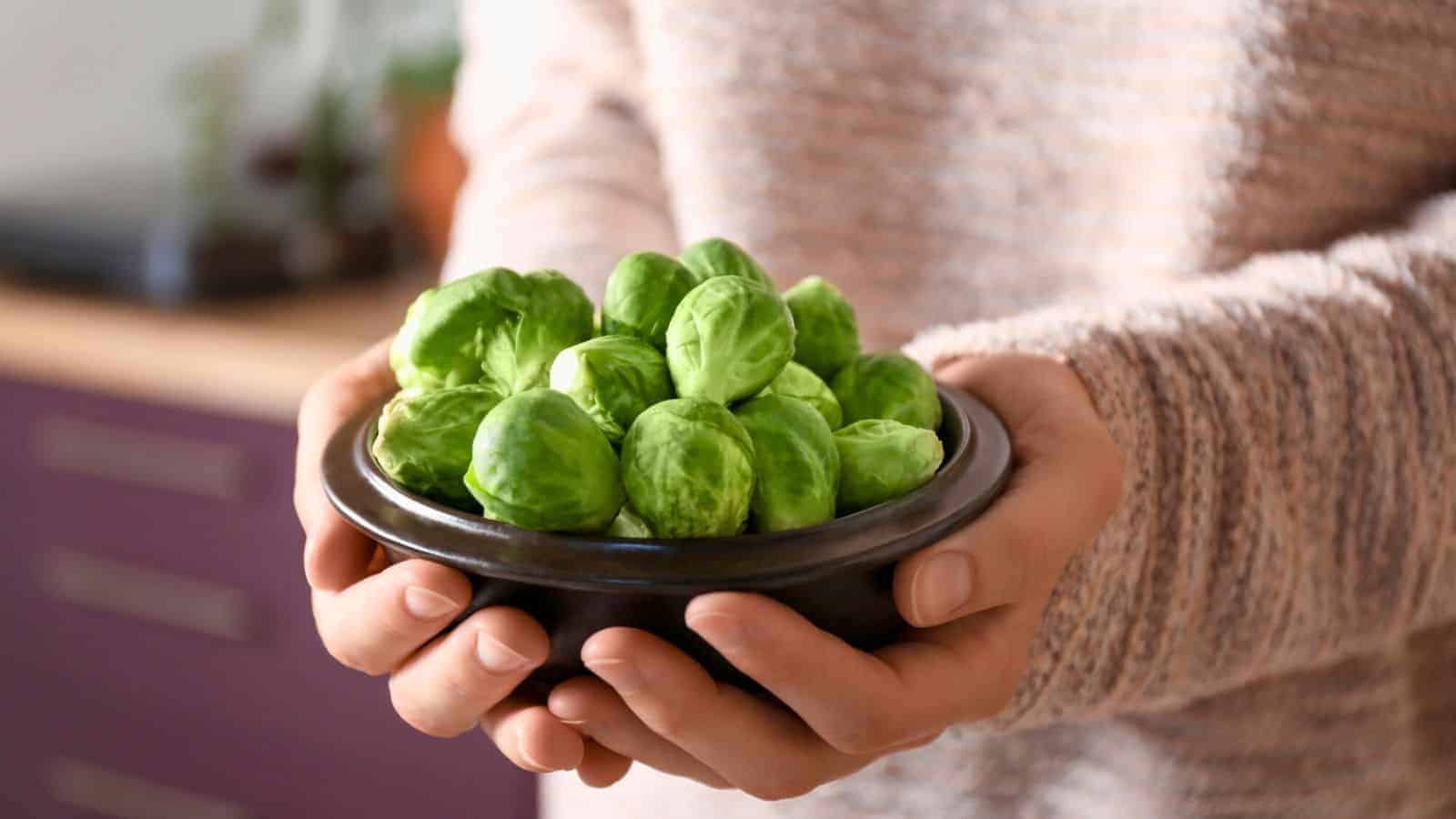 A person holding a black bowl filled with fresh Brussels sprouts, wearing a beige knitted sweater. The background is slightly out of focus, showing a kitchen setting.