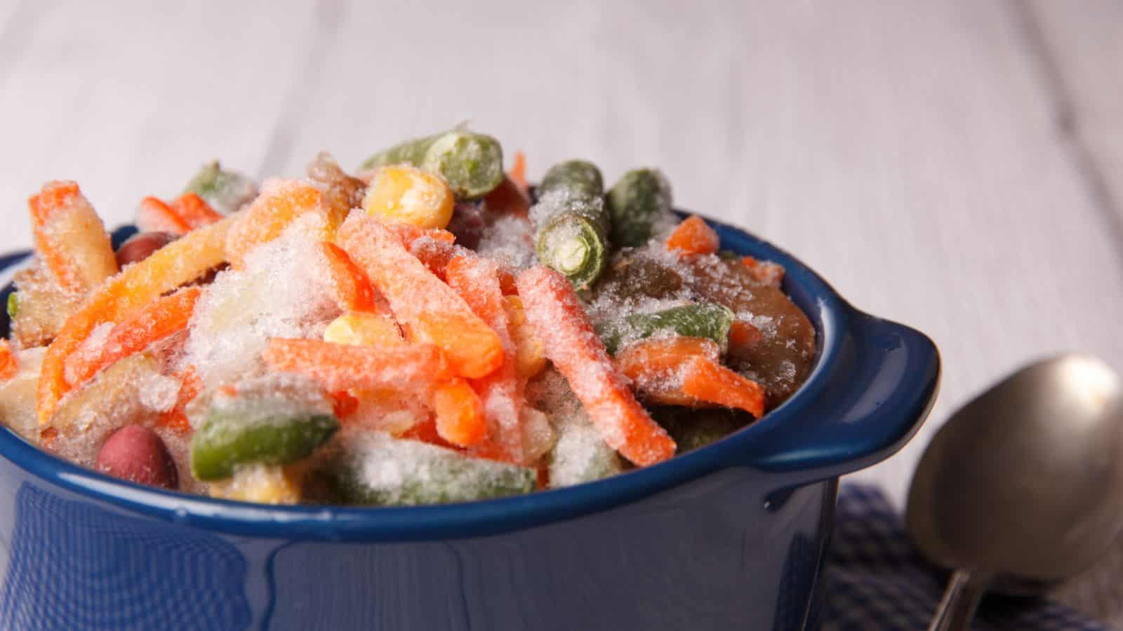 A blue bowl filled with a mix of frozen vegetables, including carrots, green beans, and corn, sits on a white wooden surface. A spoon is placed nearby on a checkered cloth.