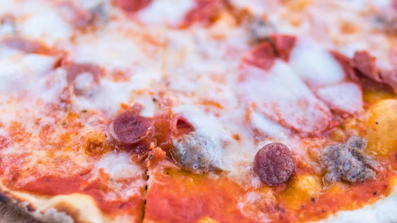 A close-up of a pizza topped with melted cheese, pepperoni slices, and sausage pieces on a thin crust. Visible tomato sauce spreads under the toppings. The edge of the pizza is slightly crispy and browned.