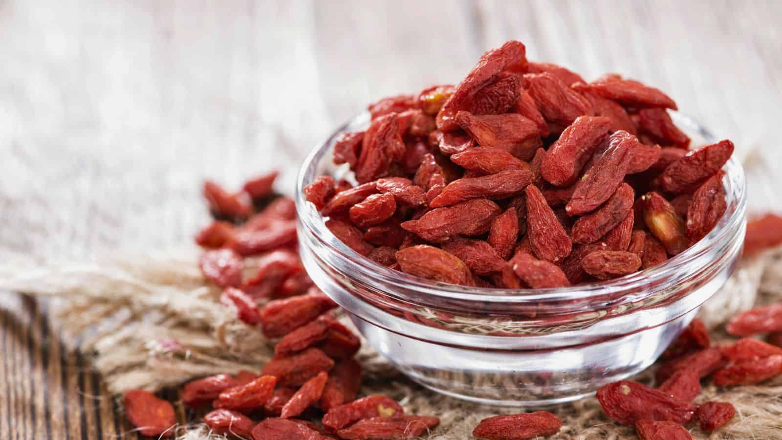 A clear glass bowl filled with dried goji berries is placed on a piece of burlap fabric on a wooden surface. Additional goji berries are scattered around the bowl. The berries have a rich red color and a slightly wrinkled texture.