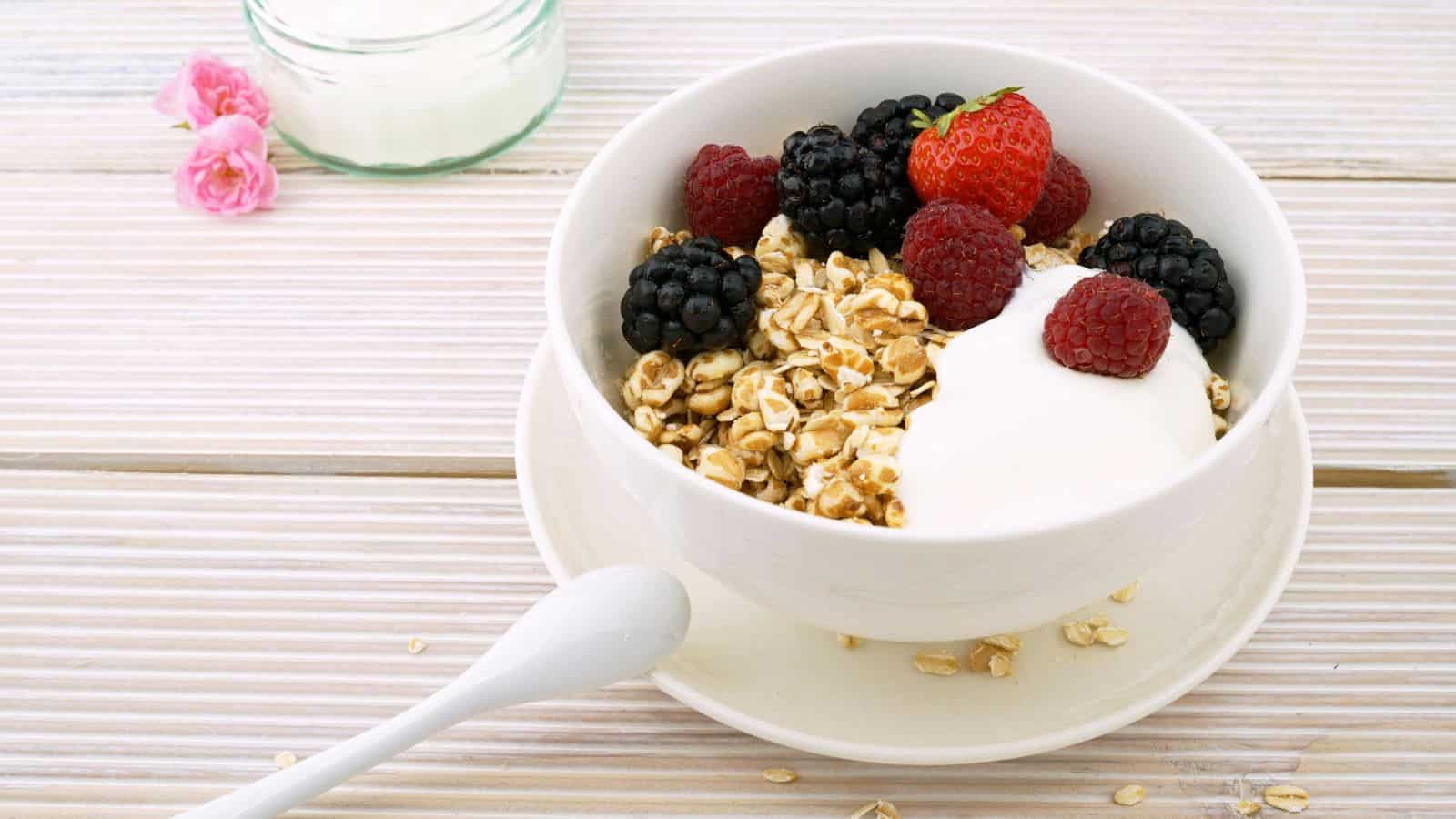 A bowl of yogurt topped with fresh blackberries, raspberries, and a strawberry, alongside a serving of granola. A spoon rests on the saucer, and a jar with a white substance is partially visible in the background, with pink flowers nearby.