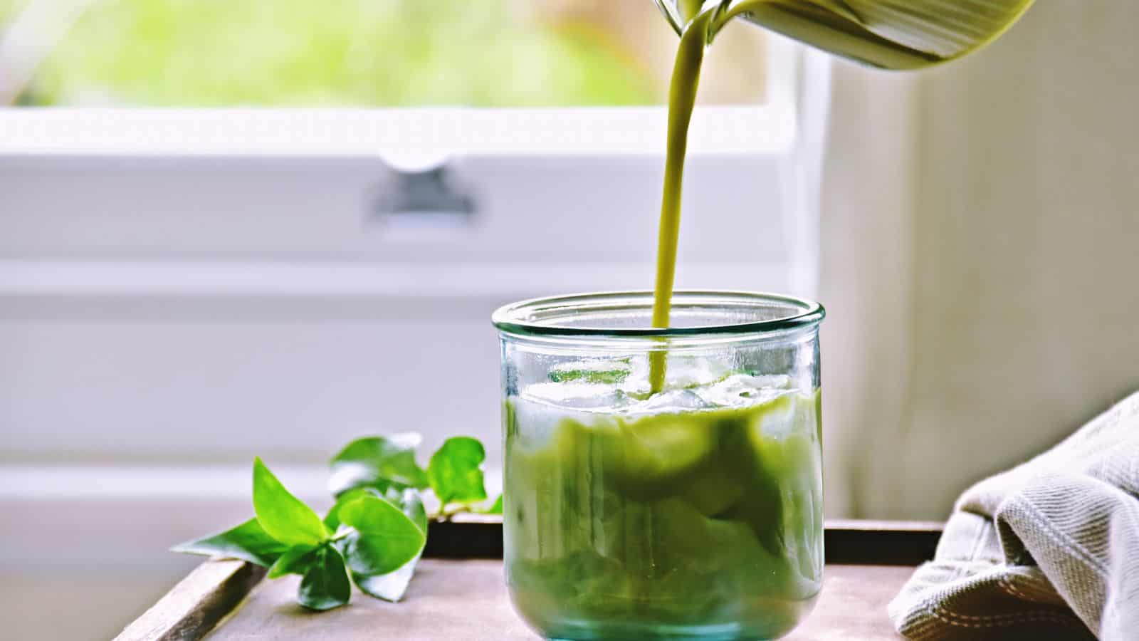 A vibrant green liquid is being poured into a glass of ice on a rustic wooden surface. Beside the glass, a small leafy plant complements the scene while natural light streams through the window, enhancing the tranquility. A neatly folded napkin is partially visible to the right, as if set by auto draft.