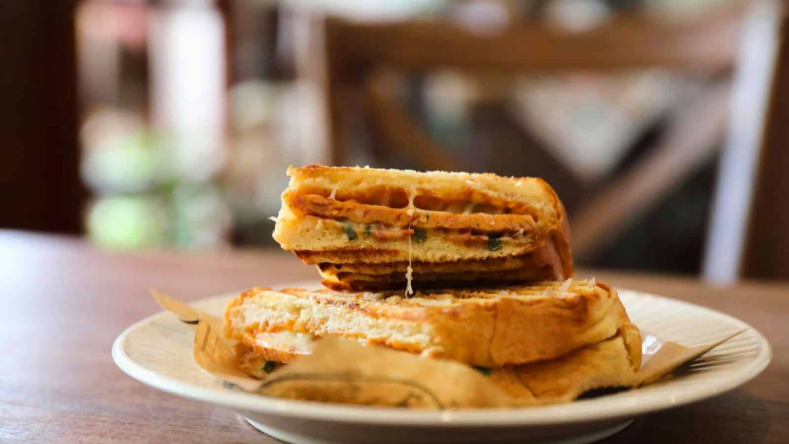A close-up of a toasted sandwich cut in half and stacked on a white plate. The sandwich filling includes melted cheese and slices of meat, with visible grill marks on the bread. The background is softly blurred.