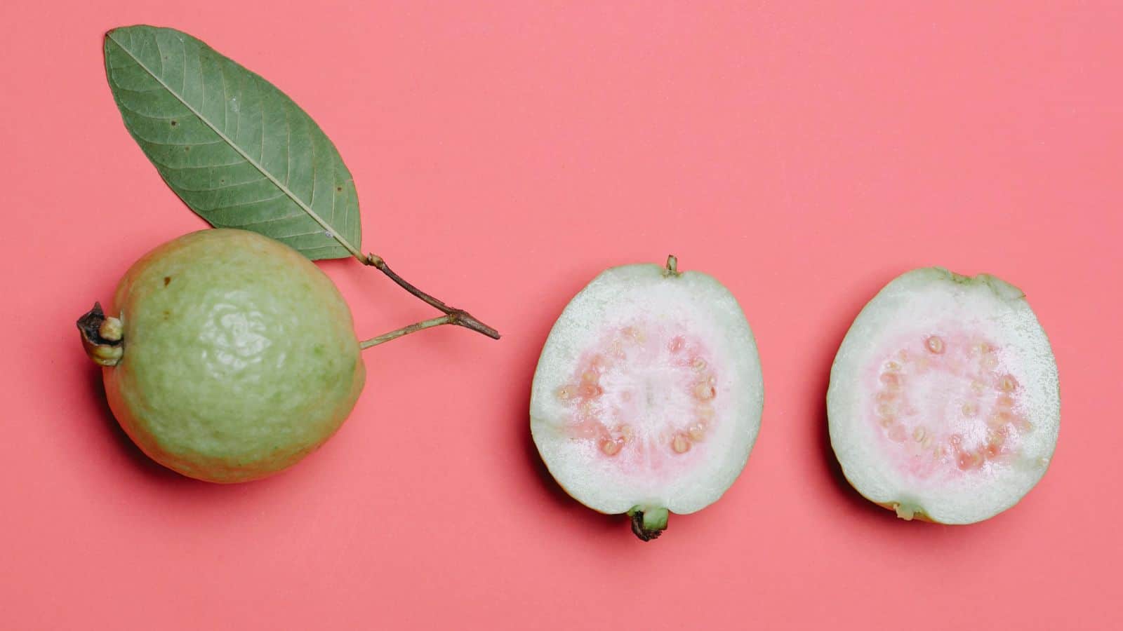 A whole guava with a leaf attached sits next to a halved guava on a pink background. This underrated fruit, often overlooked despite being nutritional powerhouses, reveals its light insides dotted with visible seeds.
