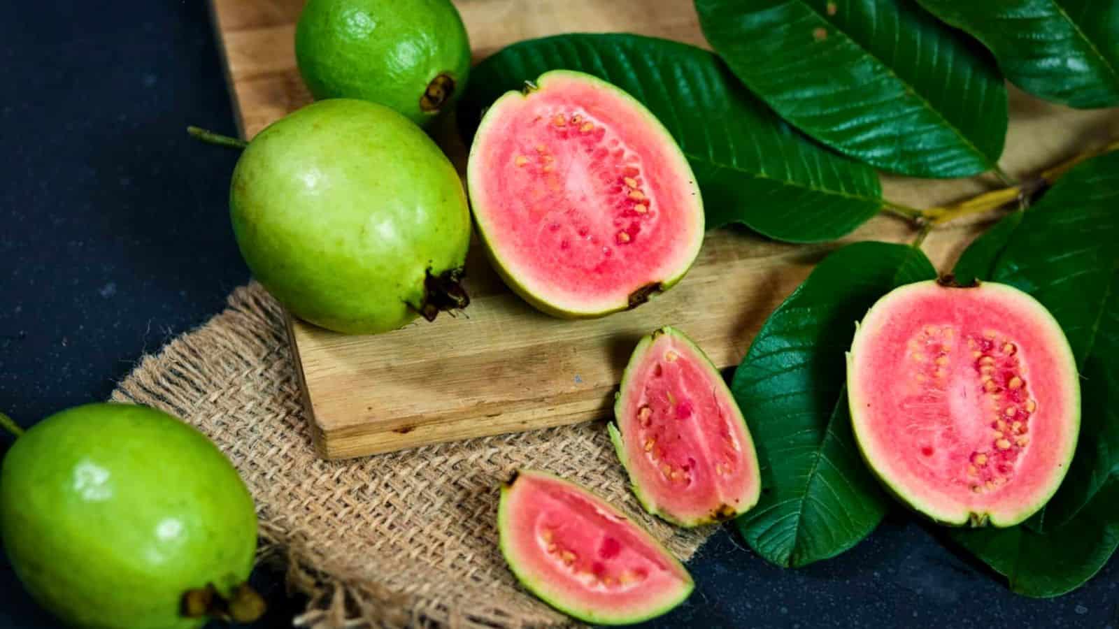 A wooden board with whole and sliced pink guavas, surrounded by green leaves. A burlap fabric partially covers the surface beneath. The guavas have a smooth green exterior and vibrant pink flesh with small seeds.