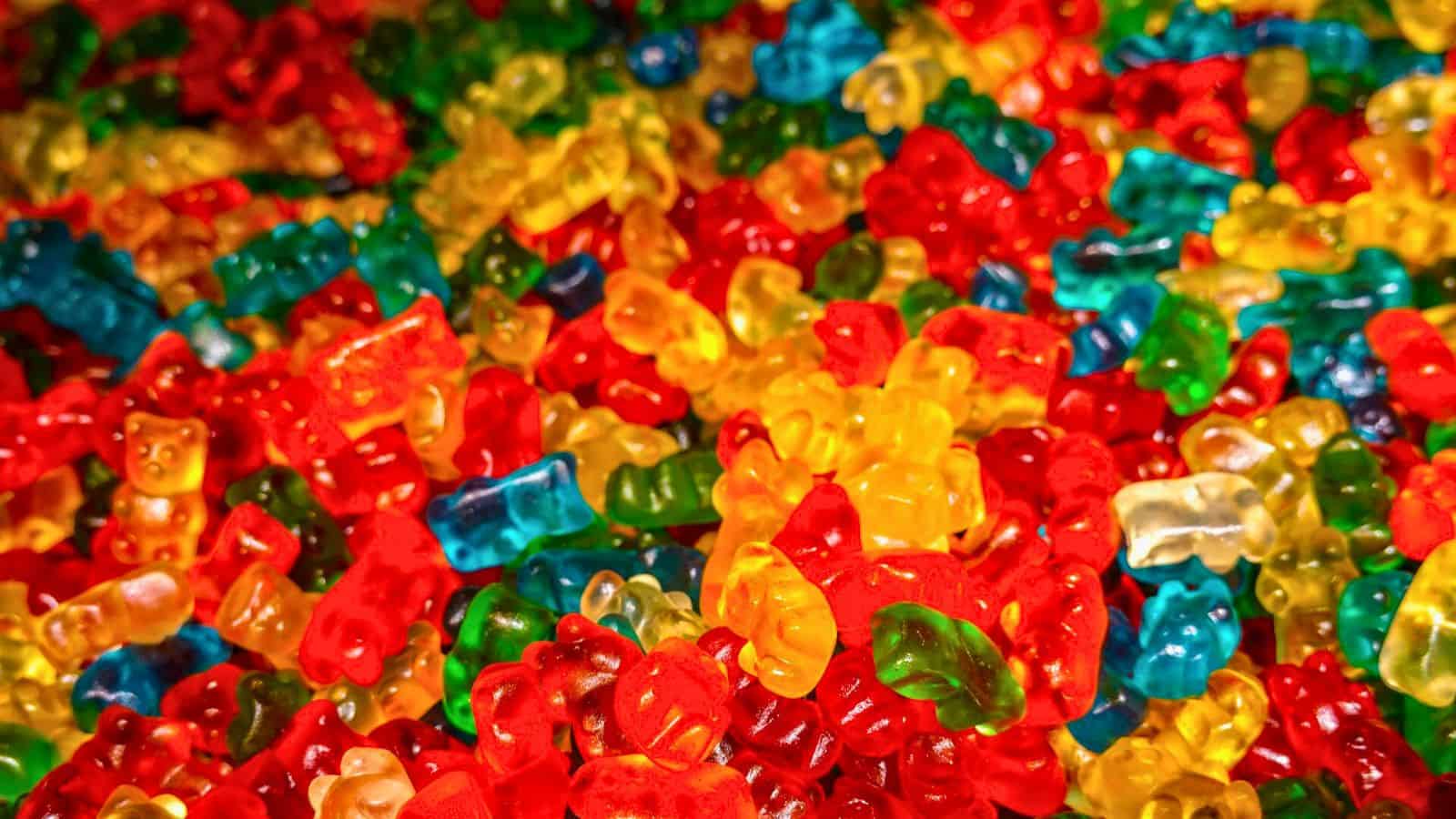 A colorful assortment of gummy bears in various shades of red, green, yellow, orange, and blue fills the frame. The candies are translucent, with some displaying mixed colors. The image is focused on the vibrant texture and variety.