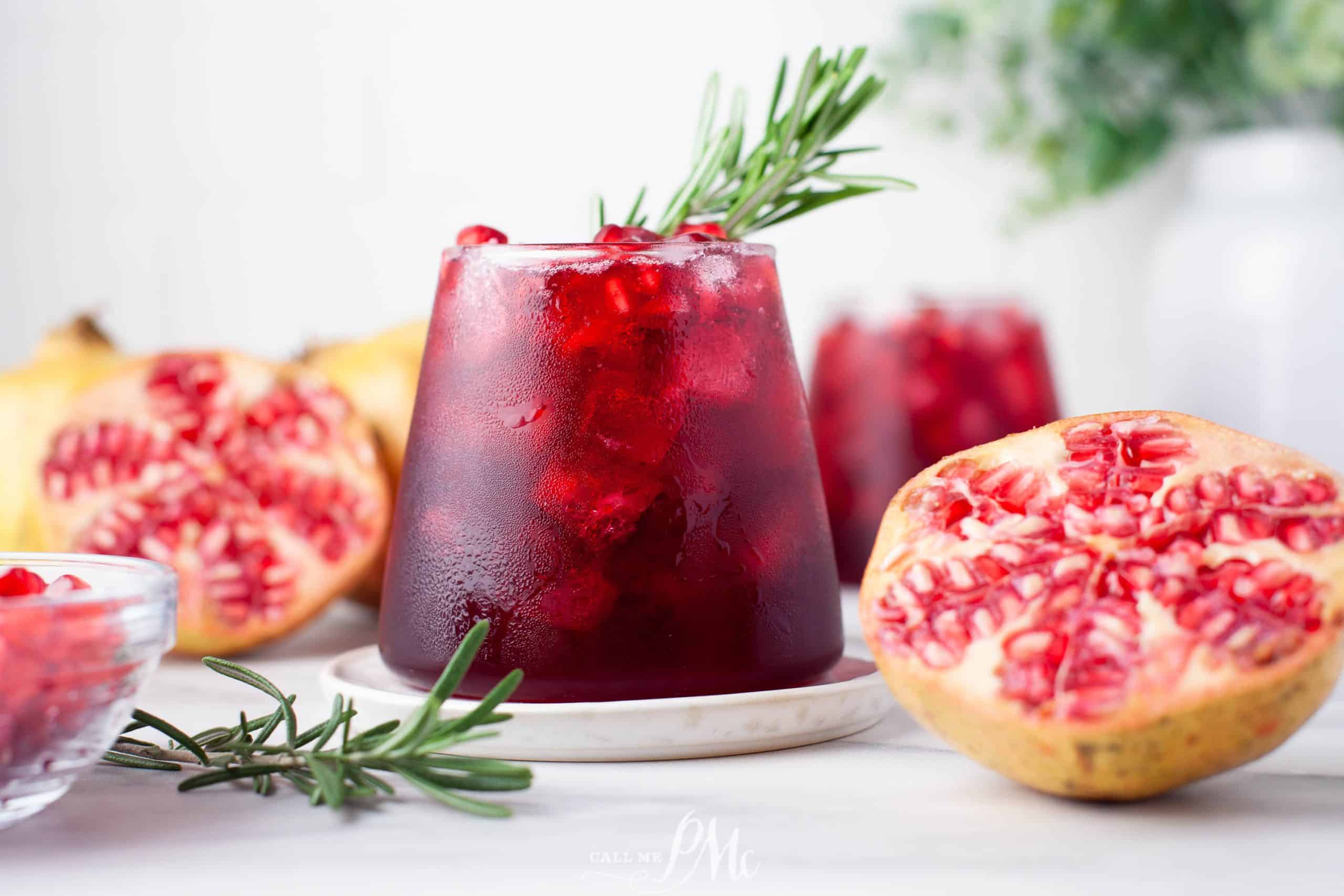 A glass filled with a dark red beverage, garnished with pomegranate seeds and a sprig of rosemary, sits on a white surface like an auto draft ready for perfection. Halved pomegranates and additional glasses containing the same drink adorn the background.