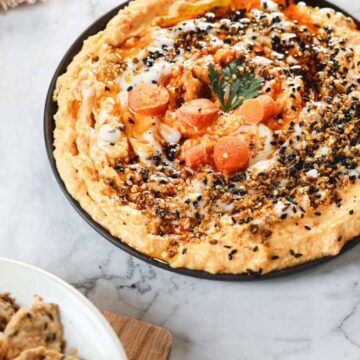 A plate of hummus, garnished with sesame seeds, spices, sliced carrots, and a sprig of parsley sits on a marble surface. A bowl of seeded crackers is nearby on a wooden board—perfect for sneaky ways to add vegetables to kids' meals.