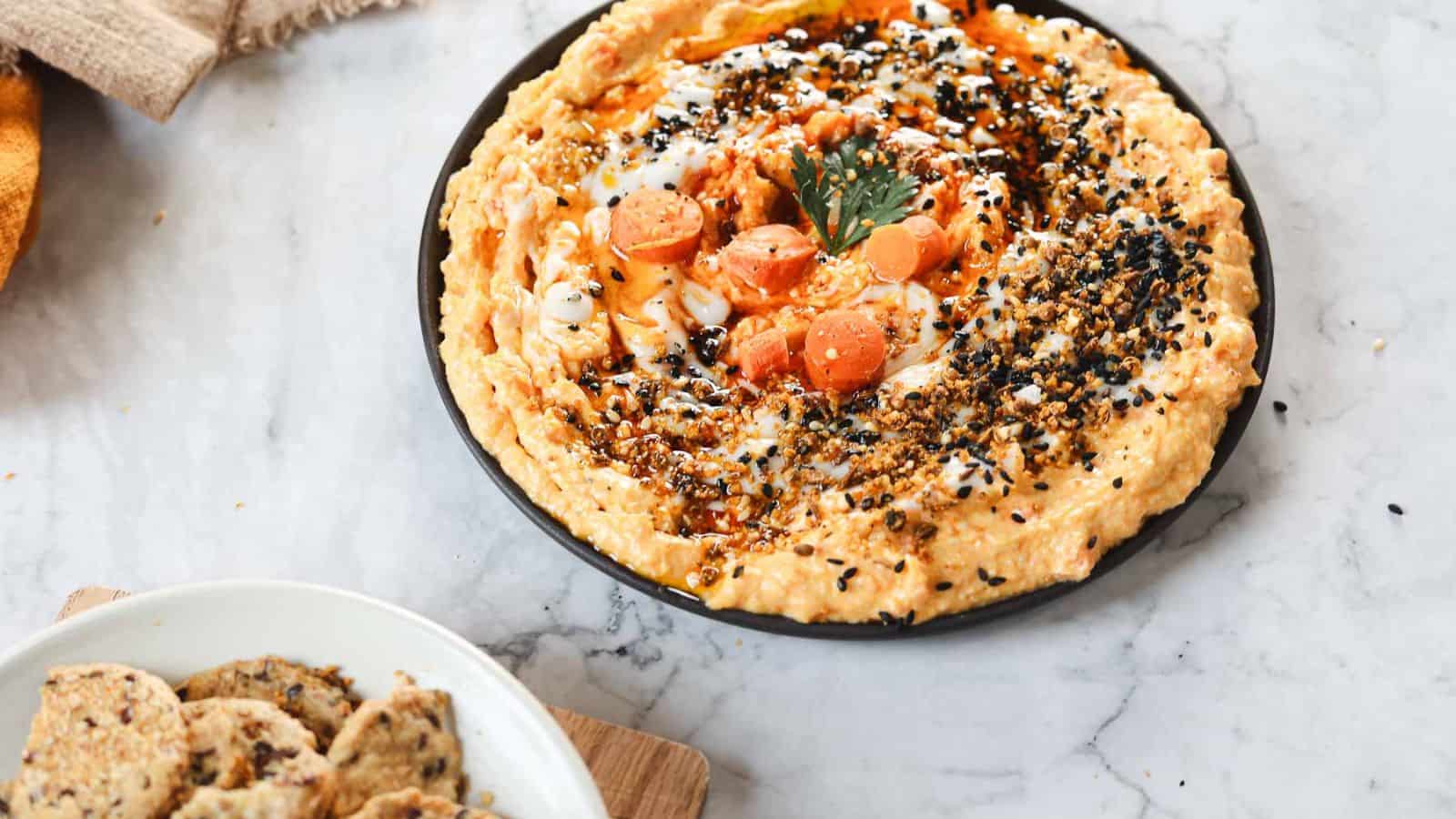 A plate of hummus, garnished with sesame seeds, spices, sliced carrots, and a sprig of parsley sits on a marble surface. A bowl of seeded crackers is nearby on a wooden board—perfect for sneaky ways to add vegetables to kids' meals.