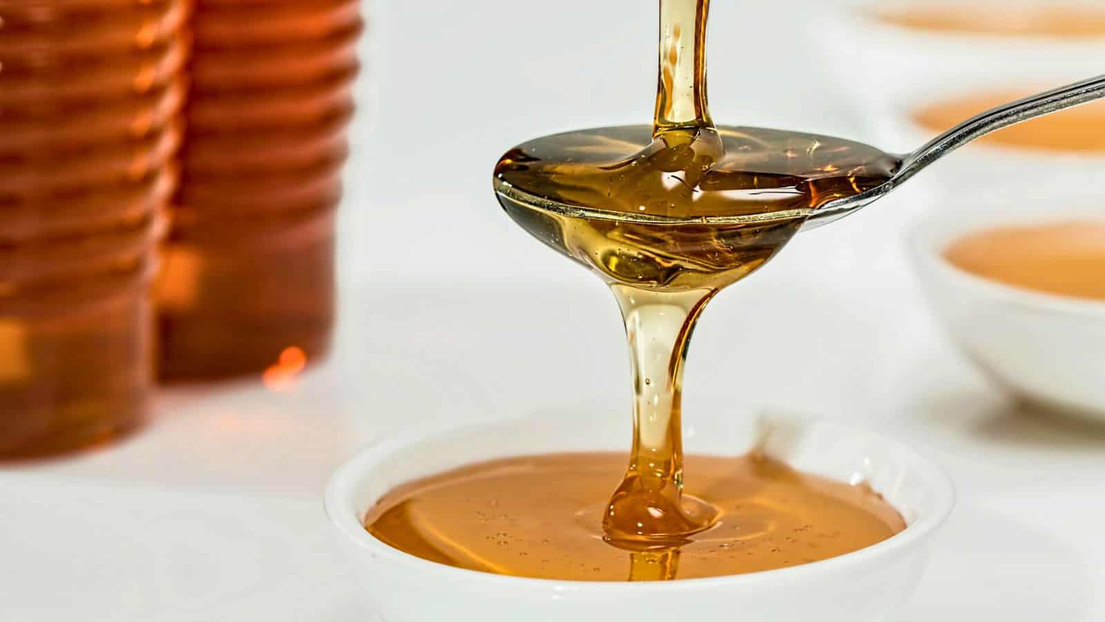 Honey being poured from a spoon into a white bowl, with bottles of honey in the background. The honey is clear and golden, creating a smooth, glossy stream.