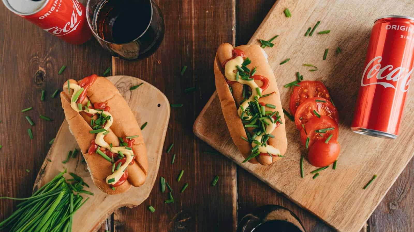 Two hot dogs topped with mustard, ketchup, and chopped chives on wooden boards. Nearby are tomato slices, a bunch of chives, a glass of dark soda, and two cans of Coca-Cola on a wooden table.