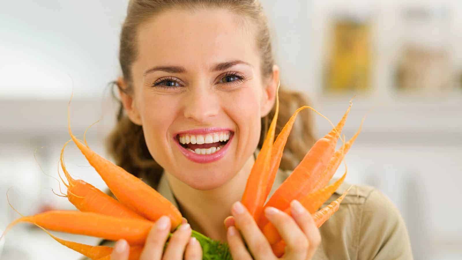 A person smiling and holding several fresh carrots with leafy green tops. The background is softly blurred, suggesting an indoor setting.