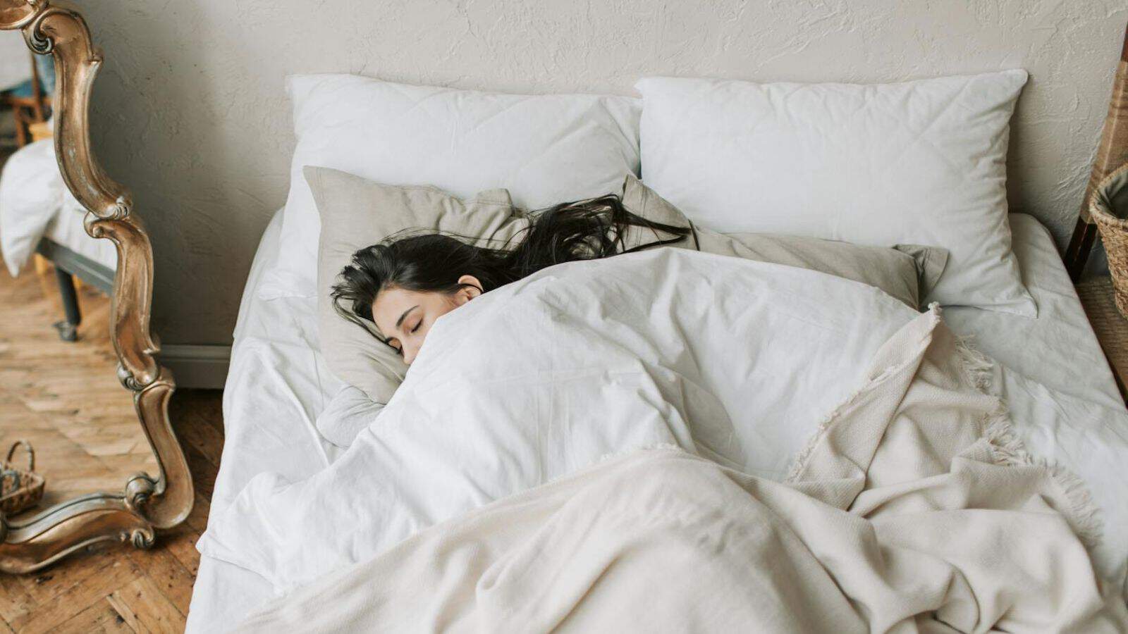 A woman with long dark hair is sleeping under white bedding on a bed with multiple pillows. The room features a wooden floor, a wicker basket, and a decorative mirror with a wooden frame next to the bed.