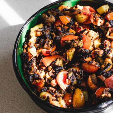 A bowl of grilled vegetables including zucchini, tomatoes, and mushrooms sits on a light-colored countertop, with sunlight casting shadows across the surface.
