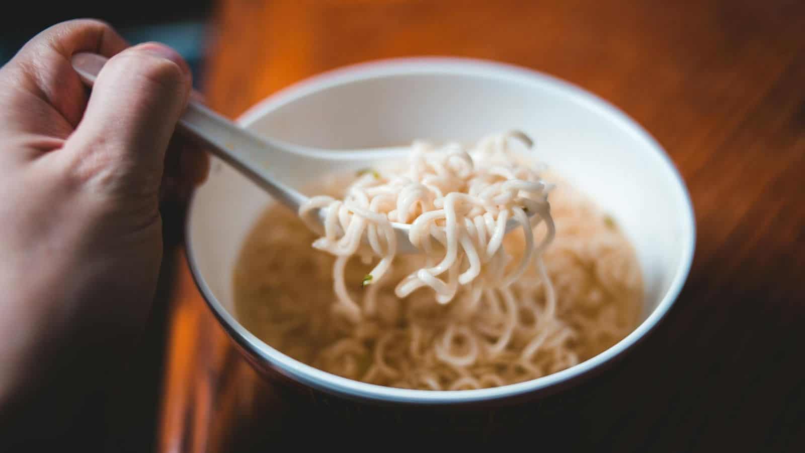 A hand holding a spoon lifts noodles from a white bowl filled with soup. The bowl rests on a wooden surface, and the soup appears clear with visible noodles.