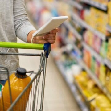 A person is pushing a shopping cart filled with groceries down an aisle in a supermarket. They are holding a tablet in one hand while resting the other on the cart handle. Various items are stocked on the shelves in the background.
