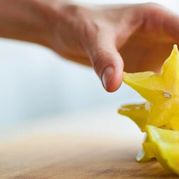 A hand reaches for a sliced starfruit on a wooden surface. The star-shaped slices are yellow and glossy, with visible seeds in the center. The background is softly blurred.