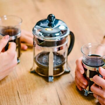 Two people sit at a wooden table, each holding a glass mug filled with coffee. A stainless steel French press sits between them, partially filled with coffee. The scene suggests a casual coffee break.