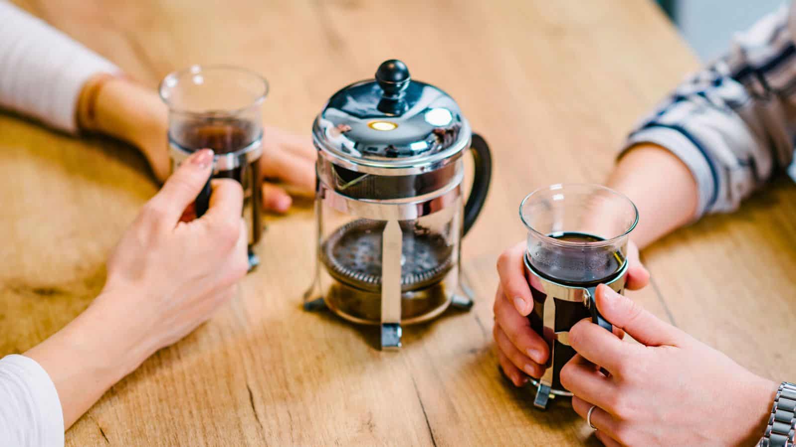 Two people sit at a wooden table, each holding a glass mug filled with coffee. A stainless steel French press sits between them, partially filled with coffee. The scene suggests a casual coffee break.