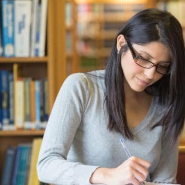 A woman with long dark hair and glasses is sitting at a desk in a library, writing in a notebook. She is wearing a gray sweater. Bookshelves filled with books are visible in the background.