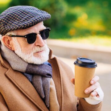 An elderly man with a white beard, wearing sunglasses, a flat cap, and a brown coat, sits outside holding a disposable coffee cup. He is also wearing a scarf and appears to be in a park setting on a sunny day.