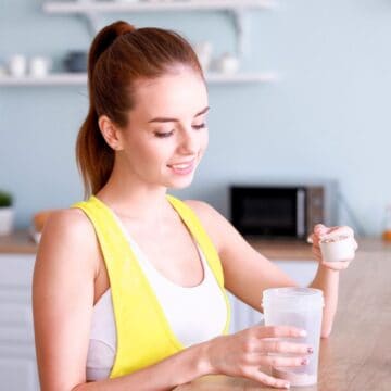 A woman in a yellow tank top is scooping protein powder into a shaker bottle in a kitchen. There are shelves with cups and a microwave in the background.
