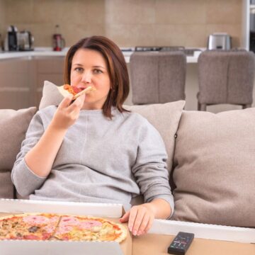 A person is sitting on a couch eating pizza from an open box. The living room has a view of the kitchen with gray chairs and a small potted plant. A remote control is placed on the couch next to them.
