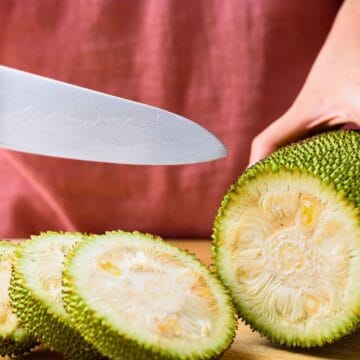 A person slices a jackfruit on a wooden cutting board with a large knife. The jackfruit has a green, spiky exterior and pale yellow interior. The person is wearing a pink shirt.