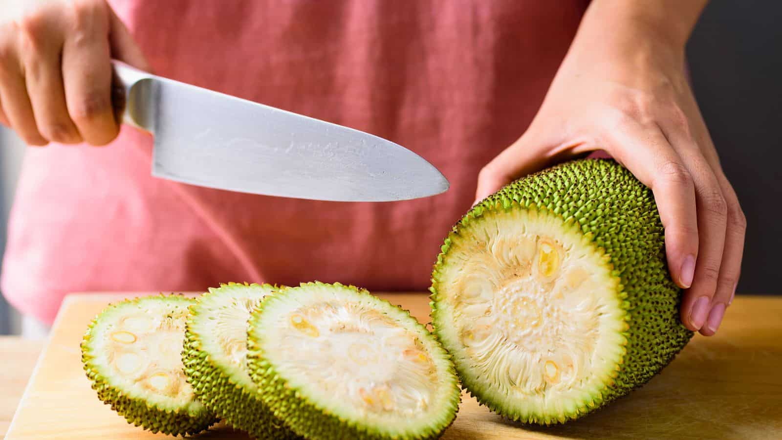 A person slices a jackfruit on a wooden cutting board with a large knife. The jackfruit has a green, spiky exterior and pale yellow interior. The person is wearing a pink shirt.