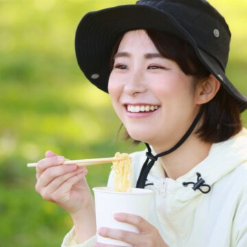A person wearing a black hat and a light jacket is smiling while holding chopsticks with noodles and a cup in an outdoor setting with a green, blurred background.