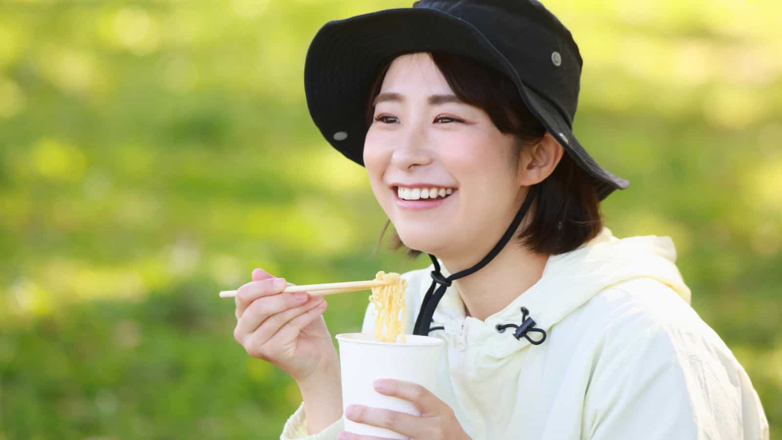 A person wearing a black hat and a light jacket is smiling while holding chopsticks with noodles and a cup in an outdoor setting with a green, blurred background.