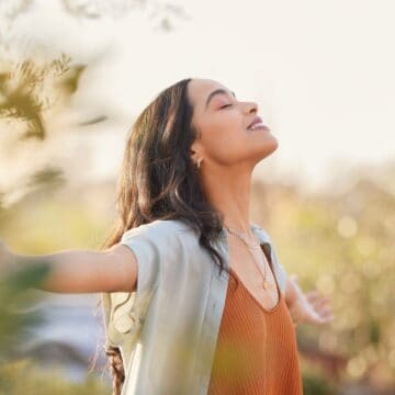 A woman with long dark hair stretches her arms out and looks upward with her eyes closed. She is standing outdoors surrounded by greenery, wearing a light blue cardigan over a rust-colored top. The lighting is bright, suggesting a sunny day.