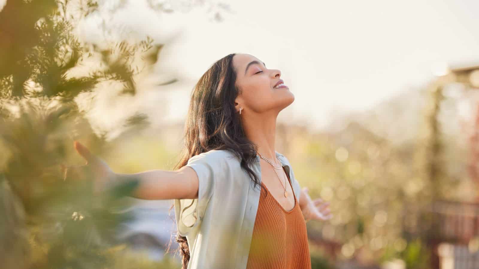 A woman with long dark hair stretches her arms out and looks upward with her eyes closed. She is standing outdoors surrounded by greenery, wearing a light blue cardigan over a rust-colored top. The lighting is bright, suggesting a sunny day.