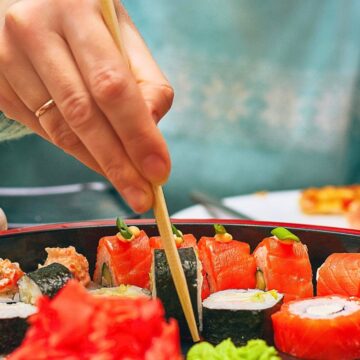 A person wearing a teal sweater uses chopsticks to pick up sushi from a platter. The sushi assortment includes various rolls and nigiri, surrounded by wasabi and ginger. A white cup is visible in the background.