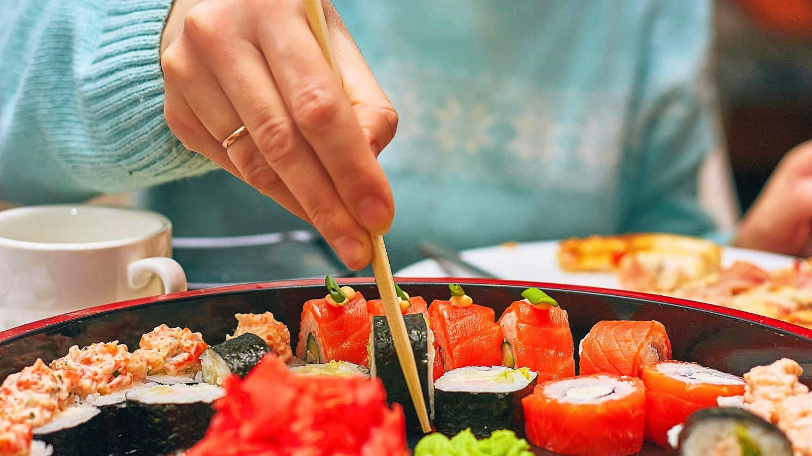 A person wearing a teal sweater uses chopsticks to pick up sushi from a platter. The sushi assortment includes various rolls and nigiri, surrounded by wasabi and ginger. A white cup is visible in the background.