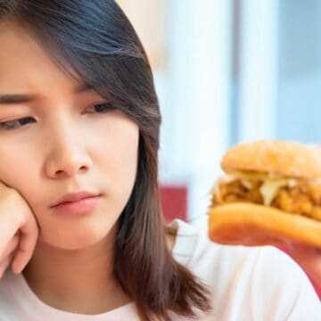 A person gazes thoughtfully at a sandwich being offered by another individual. The sandwich, with its bread bun and visible filling, seems to hold a decision in question. In an indoor setting with a blurred background, the scene subtly hints at fast food to avoid, sparking hesitation.