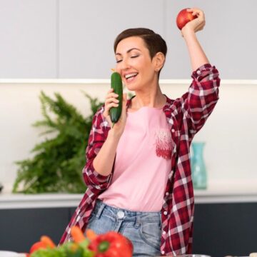 A person in a red plaid shirt holds a cucumber and an apple in a kitchen. Various vegetables are on the counter, with a leafy plant in the background. The person appears to be singing or speaking into the cucumber.
