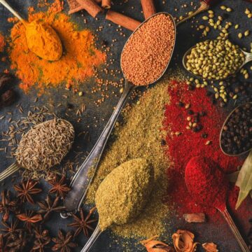 An assortment of spices displayed on a dark surface with vintage spoons. Spices include turmeric, red lentils, coriander seeds, cumin, red chili powder, black peppercorns, and star anise, alongside bay leaves and cinnamon sticks.