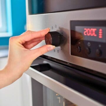 A hand adjusts the temperature dial on an oven, set to 200°C. The oven has a digital display and is located in a modern kitchen with bright blue walls and a window in the background.