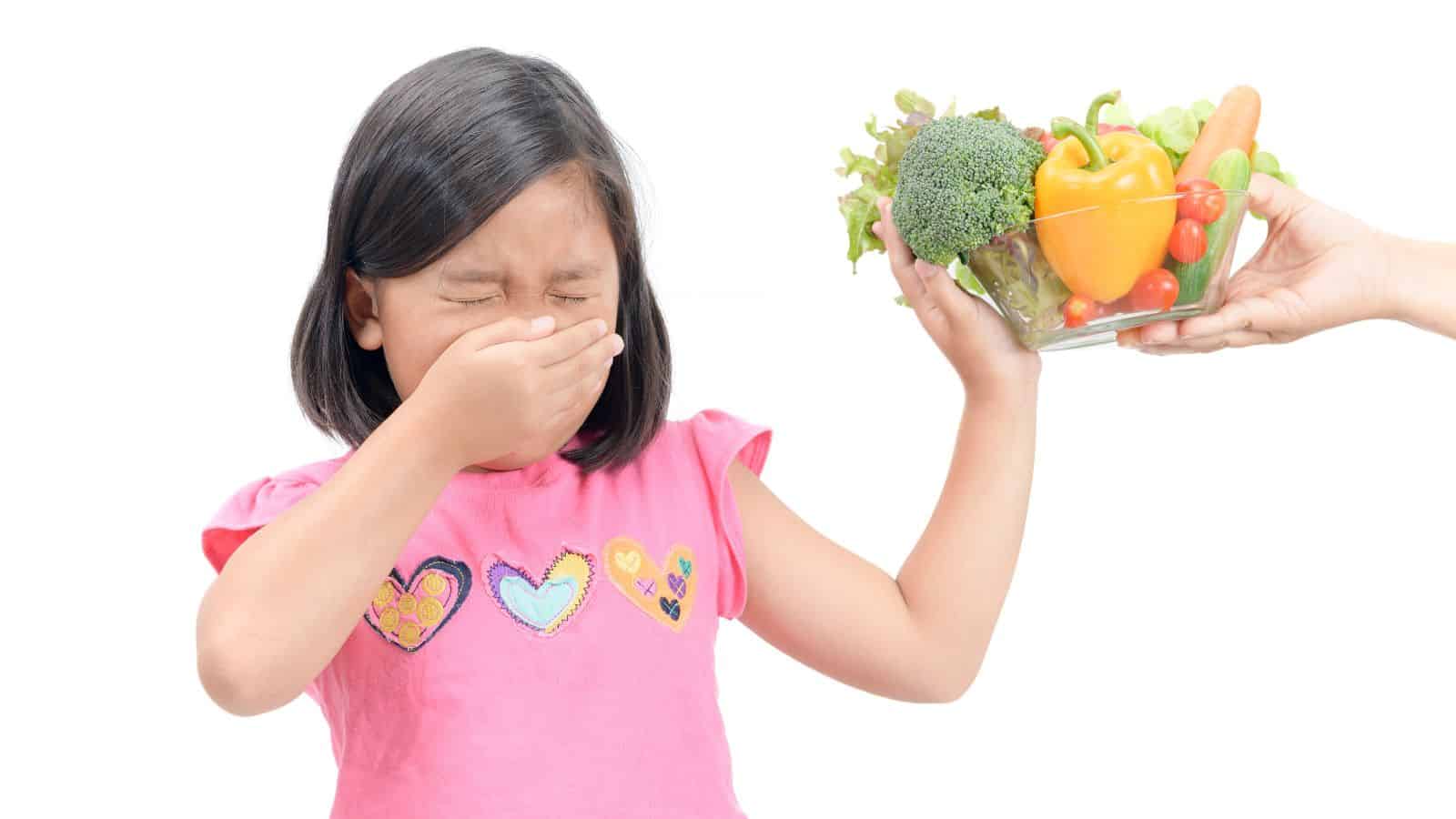 A girl in a pink shirt holding her nose while another hand offers her a bowl of vegetables, including broccoli, bell pepper, cherry tomatoes, and lettuce. The girl's eyes are closed, and she appears to be reacting to the vegetables.