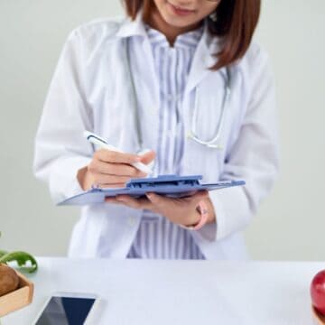 A person in a white lab coat writes on a clipboard. They are standing behind a table with various fruits and vegetables, including apples, lemons, and greens. A smartphone is also visible on the table.