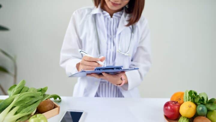 A person in a white lab coat writes on a clipboard. They are standing behind a table with various fruits and vegetables, including apples, lemons, and greens. A smartphone is also visible on the table.